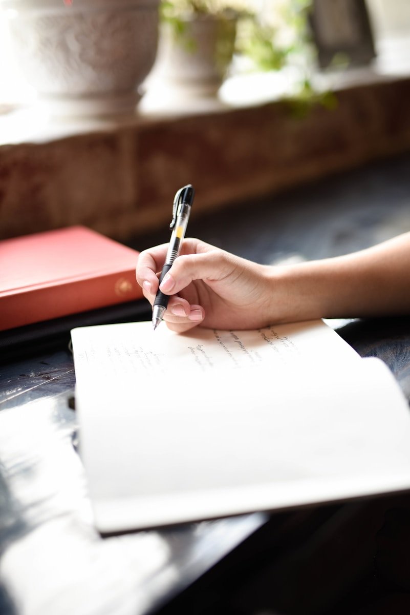 Main écrivant avec un stylo sur papier sur une table, livre et plantes derrière.