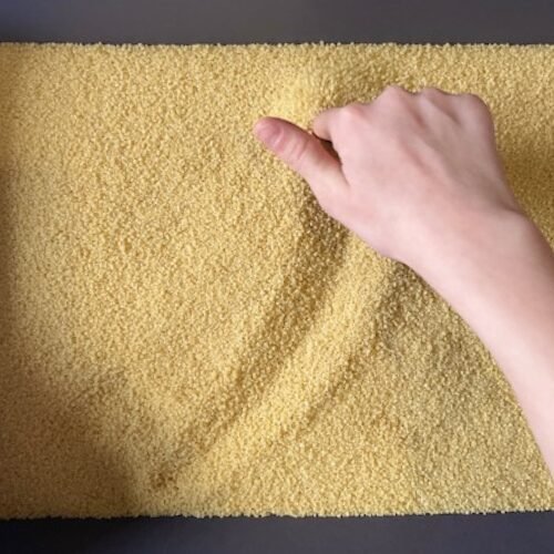 Main traçant un sillon dans des grains de semoule ou couscous sur une surface plate.