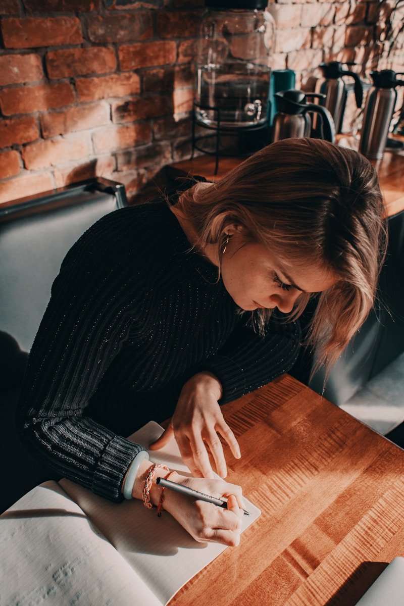 Personne concentrée écrivant dans un cahier sur une table en bois devant un mur en briques.